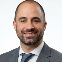 Portrait photograph of a person with short hair and a beard wearing a grey suit, white shirt and tie against a white background