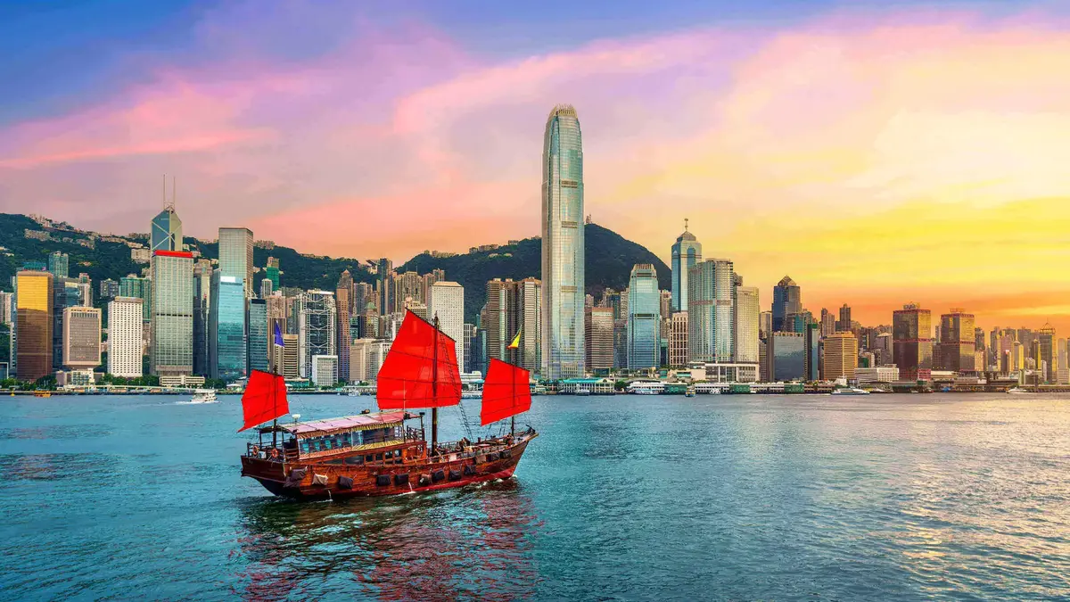 Hongkong_Skyline_Junkboot_Sonnenuntergang Traditionelles chinesisches Junkboot mit roten Segeln fährt vor der Skyline von Hongkong bei farbenprächtigem Sonnenuntergang über das Wasser.